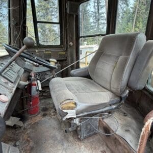 24 4 - Montana Auctions by Kevin Hill Interior of an old, dusty tractor cab with a torn driver's seat, rusty floor, and a red fire extinguisher mounted near the wheel.