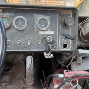 24 7 - Montana Auctions by Kevin Hill Close-up of a dusty tractor dashboard with round gauges, ignition switch, and exposed wiring behind a weathered panel. key in the ignition with tags nearby.