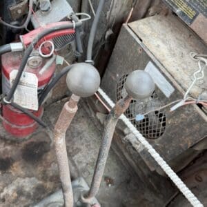 24 9 - Montana Auctions by Kevin Hill Two rusted gear shift levers with black spherical knobs in an old, dusty machine; a red fire extinguisher and rusted panels nearby.