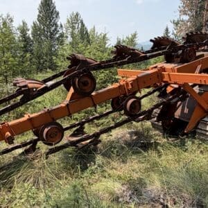 25 1 - Montana Auctions by Kevin Hill Decommissioned orange agricultural header resting on dry grass beside a forested area, rusted and abandoned.
