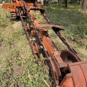 25 2 - Montana Auctions by Kevin Hill Old rusty trenching machine with chained digging blades in a grassy clearing.