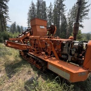 25 3 - Montana Auctions by Kevin Hill Abandoned orange tracked forestry machine in a pine forest, with exposed mechanical arms and rusted metal.