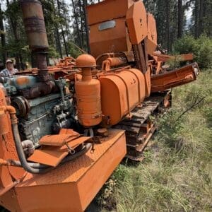 25 5 - Montana Auctions by Kevin Hill Old orange industrial machine in a forest, with rusted components and tracks.