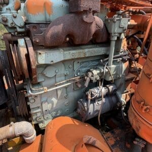 25 6 - Montana Auctions by Kevin Hill Rusty industrial diesel engine with turquoise housing and a large rusted exhaust manifold, surrounded by orange machinery outdoors in a wooded area