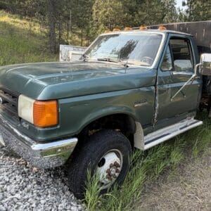 26 1 - Montana Auctions by Kevin Hill Old green Ford dump truck parked on gravel beside a grassy field, showing rust and wear.