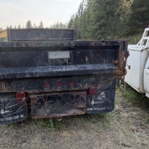 26 2 - Montana Auctions by Kevin Hill Rear view of a rusty dump truck with weathered mud flaps and a faded logo, parked beside a white utility truck; construction equipment and trees in the background.
