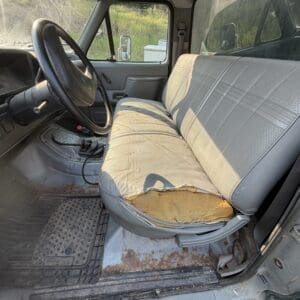 26 2 - Montana Auctions by Kevin Hill Interior of an old pickup truck with a torn bench seat, dusty floor, and a worn steering wheel visible from the driver’s side, daylight seeping through the windows.