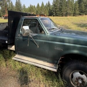 26 3 - Montana Auctions by Kevin Hill Green vintage pickup with a flatbed parked in tall grass beside a gravel area, with a forested backdrop and construction equipment in the distance.
