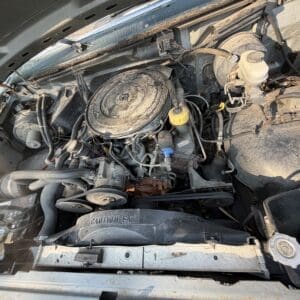 26 5 - Montana Auctions by Kevin Hill Dusty vehicle engine bay with belts, hoses, and an round air filter housing at center; radiator shroud reads 'CAUTION FAN'.
