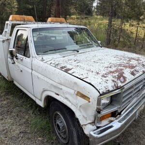 27 2 - Montana Auctions by Kevin Hill White rusty utility truck with orange lights parked in a grassy forest area.