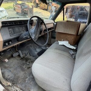27 2 - Montana Auctions by Kevin Hill Interior of an old truck with a dusty dashboard and steering wheel; cardboard boxes sit on the passenger bench and a muddy floor shows wear in a junkyard setting.