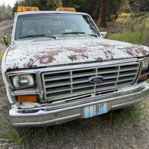 27 3 - Montana Auctions by Kevin Hill Front view of a white Ford pickup with heavy rust and peeling paint, chrome bumper, and orange emergency lights on top, parked on a dirt field near trees.