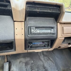 27 4 - Montana Auctions by Kevin Hill Old tan car dashboard center panel with a 'CUSTOM' label, aftermarket cassette stereo, and a lower radio unit; dusty and worn interior.