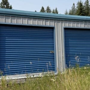 28 1 - Montana Auctions by Kevin Hill Blue corrugated metal storage units with two large blue roll-up doors, surrounded by tall grass and trees under a clear sky in a rural lot.