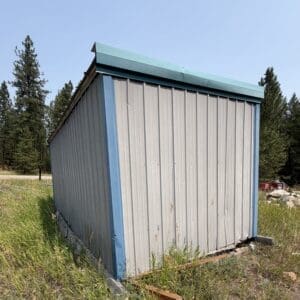 28 2 - Montana Auctions by Kevin Hill Metal storage shed with blue trim and teal roof, standing in a grassy rural area with trees in the background.