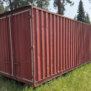 29 1 - Montana Auctions by Kevin Hill Red shipping container on a grassy lot with trees in the background, viewed from the side.