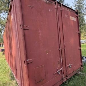 29 2 - Montana Auctions by Kevin Hill Red rusted metal shipping container on a grassy field with trees in the background.