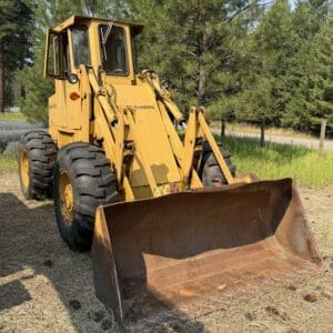 3 1 - Montana Auctions by Kevin Hill Yellow front-end loader with a large rusted bucket parked on dry grass, near a road with pine trees in the background.
