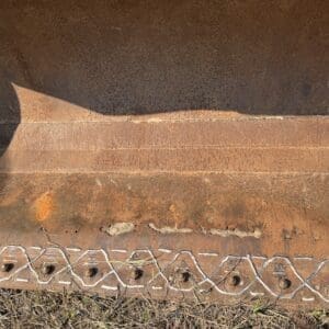 3 1 - Montana Auctions by Kevin Hill Close-up of a rusted metal surface with a row of circular holes and a white zigzag line along the bottom edge, laid on dry grass.