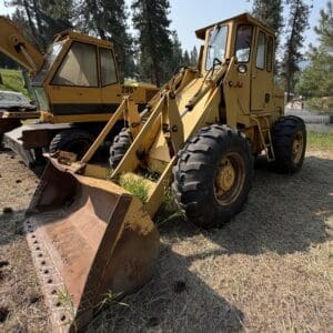 3 2 - Montana Auctions by Kevin Hill Yellow backhoe loader with a large rusted front bucket in a grassy yard, surrounded by other heavy equipment and tall trees.
