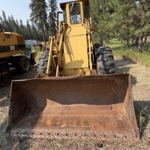 3 3 - Montana Auctions by Kevin Hill Yellow Allis-Chalmers front-end loader with a wide rusted bucket in a outdoor equipment yard, trees in the background.