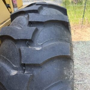 3 3 - Montana Auctions by Kevin Hill Close-up of a large rugged tractor tire with deep, chunky tread blocks on yellow machinery; fence and outdoor ground visible in the background.
