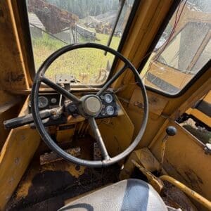 3 4 - Montana Auctions by Kevin Hill Interior of a yellow bulldozer cab with a large steering wheel, dusty floor, and worn seat; gauges on the dashboard and sunlight coming through dirty windows with yard equipment outside.