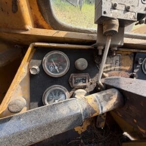 3 5 - Montana Auctions by Kevin Hill Close-up of a dusty yellow machine dashboard showing oil pressure and oil temperature gauges with a worn control lever nearby.