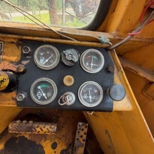 3 7 - Montana Auctions by Kevin Hill Dashboard of an old, weathered construction vehicle with five round gauges and a yellow exterior. Outside scene visible through window, dusty interior.