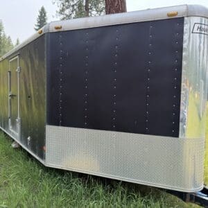 30 1 - Montana Auctions by Kevin Hill Black enclosed cargo trailer with diamond-plate front, parked on grass in a field behind a hitch edge.