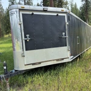 30 1 - Montana Auctions by Kevin Hill Black enclosed trailer on a dual-axle hitch parked in a grassy field with trees in the background.