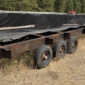 31 1 - Montana Auctions by Kevin Hill Rusty flatbed trailer with six wheels carrying long black-wrapped boards on a dry grassy field.