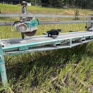 32 1 - Montana Auctions by Kevin Hill Industrial metal-cutting saw mounted on a portable metal worktable in a grassy field; weathered teal frame with a circular blade guard.