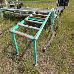 32 2 - Montana Auctions by Kevin Hill Green metal farm machine with multiple rusted rollers on a grassy field; a control panel sits to the right with cables, indicating a conveyor/cedar logging tool.