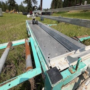 32 4 - Montana Auctions by Kevin Hill Close-up of a weathered metal trailer with rusted rails and perforated decking in a grassy field, construction vehicles in the distance.