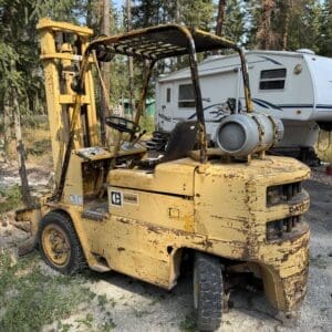 33 1 - Montana Auctions by Kevin Hill Worn yellow forklift parked outdoors beside a white travel trailer, propane tank mounted on the rear, trees in the background, dusty ground nearby.