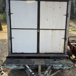 34 1 - Montana Auctions by Kevin Hill Front view of an old utility trailer with a large, four-panel metal cabinet mounted at the back, secured by a padlock and chains, in a dry field.