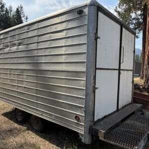 34 1 - Montana Auctions by Kevin Hill Silver aluminum utility trailer with riveted panels on a dirt lot, surrounded by pine trees and a clear sky.