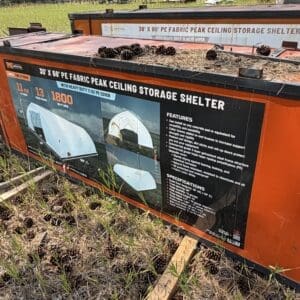 35 3 - Montana Auctions by Kevin Hill Orange fabric storage shelter with a sign reading “30' x 60' PE Fabric Peak Ceiling Storage Shelter” outdoors, with pinecones and tall grass nearby