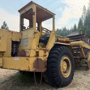 4 1 - Montana Auctions by Kevin Hill Rusty yellow scraper with a cabin and oversized rear wheel parked on a grassy field, forest in the background.