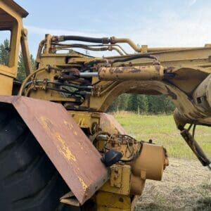 4 12 - Montana Auctions by Kevin Hill Rusty yellow construction vehicle (bulldozer/tractor) with exposed hydraulic arms in a grassy field, showing wear and rust.