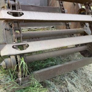 4 7 - Montana Auctions by Kevin Hill Rusty cut‑bundle or conveyor mechanism of an old farm implement with multiple metal rollers and belts, lying on grass.