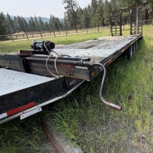 45 1 - Montana Auctions by Kevin Hill Flatbed trailer with a winch on the side, parked on tall grass in a rural field; wood deck and rear ramp folded up toward the end fence line.