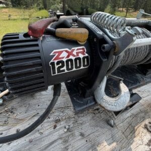 45 2 - Montana Auctions by Kevin Hill Electric winch with steel cable and forged hook on a wooden surface outdoors (ZXR 12000).