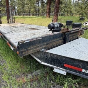 45 3 - Montana Auctions by Kevin Hill Flatbed trailer with a metal ramp in a grassy field, trees in the background and spare tires nearby.