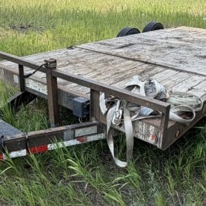 46 1 - Montana Auctions by Kevin Hill Rusty flatbed trailer with a wooden deck parked in tall grass; hitch and straps draped over the side.