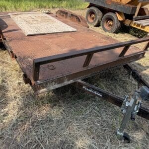 48 1 - Montana Auctions by Kevin Hill Rusty flatbed trailer in a dry grassy field, with a weathered diamond-plate deck and a yellow industrial vehicle nearby.