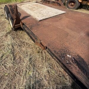 48 2 - Montana Auctions by Kevin Hill Rusty flatbed trailer with a diamond-plate deck on a dry field; a weathered plywood panel rests on top near old wheels.