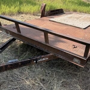 48 3 - Montana Auctions by Kevin Hill Rusty flatbed trailer on a grassy field, with hitch and jack at the front.