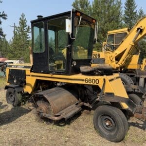 5 1 - Montana Auctions by Kevin Hill Yellow road roller with a front roller and sweeping broom in a grassy lot, machine marked 6600 and MAXI SWEEP, with another yellow excavator in the background.